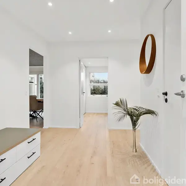 En lys entré med et hvidt skab, hvorpå der står en plante. Rundt spejl på væggen. Åben dør fører ind til et værelse med store vinduer.