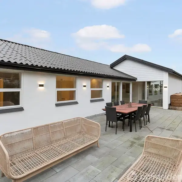 Et moderne hus med hvide vægge og sorte tagsten. Foran huset står et havemøbelsæt med borde og stole på en stenterrasse, omgivet af buske og en klar himmel.