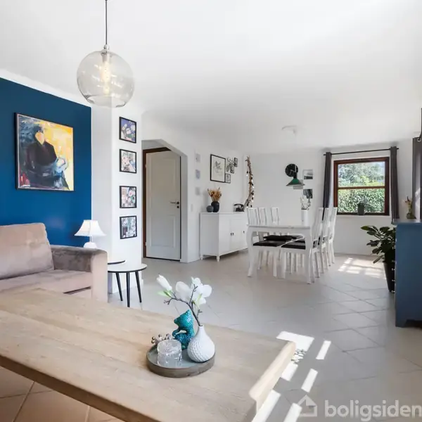 Sofa står stille ved blå væg dekoreret med kunst, mens spisebord ses i baggrunden i et lyst rum med vinduer og potteplanter.