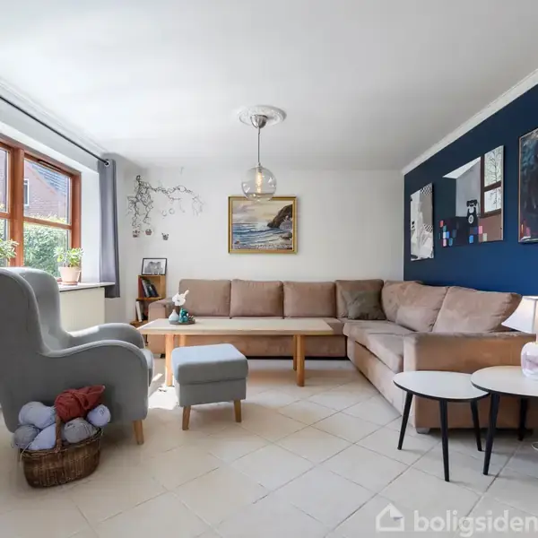 En lys stue med en lys beige hjørnesofa mod en mørkeblå væg, dekoreret med billeder. En grå lænestol står ved vinduet; en kurv med garn ved siden.