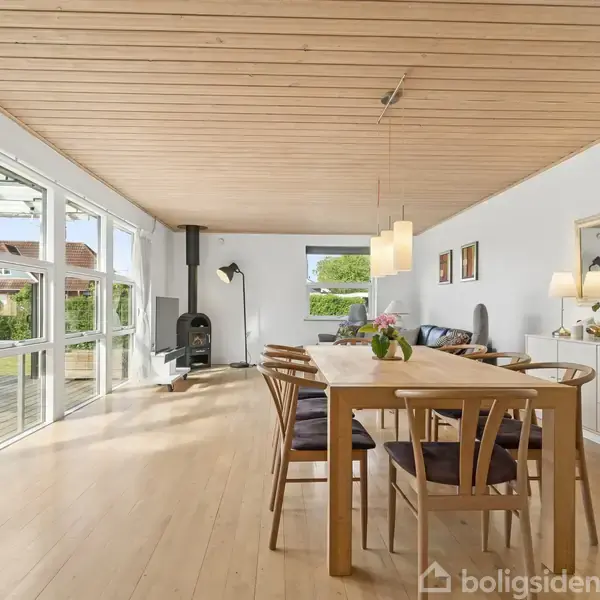 Et spisebord med stole står i et lyst rum med trægulve og loft, ved siden af en stor vinduesvæg. En brændeovn er placeret bagerst, med udsigt til en have.