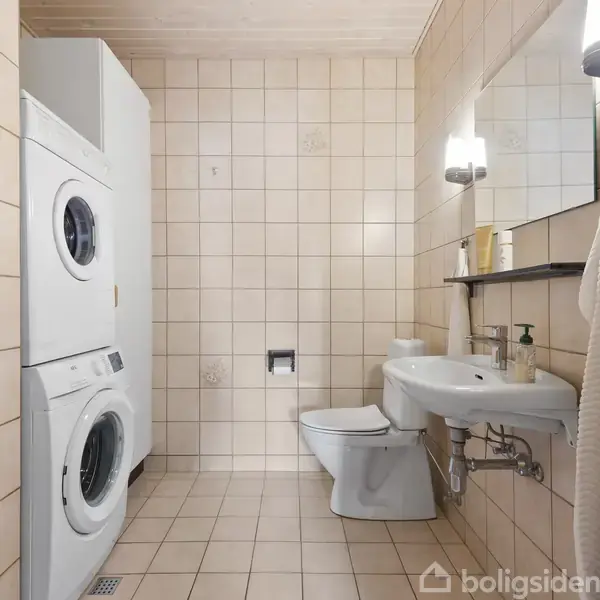 Vaskemaskine og tørretumbler står stablet i hjørnet af et flisebelagt badeværelse. Ved siden af er der en håndvask, toilet og spejl. Håndklæder hænger på væggen.