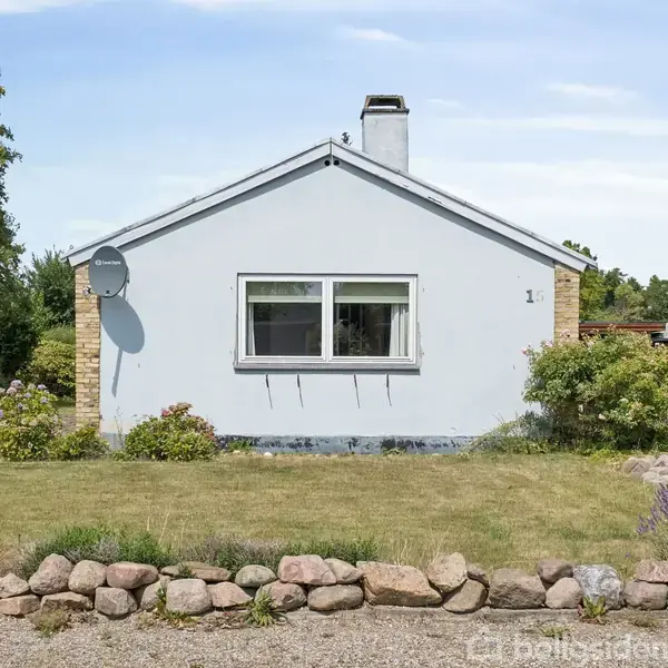 En hvid bungalow står stille, omkranset af en velplejet have med buske og lavendler. En stensætning markerer skellet mod en grusbelagt forhave.