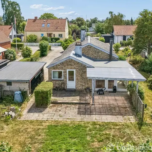 Et hus med stentag står i en villahave, flankeret af en garage. Forhaven har et flisebelagt område omgivet af grønne buske. Flere huse ses i kvarterets baggrund.