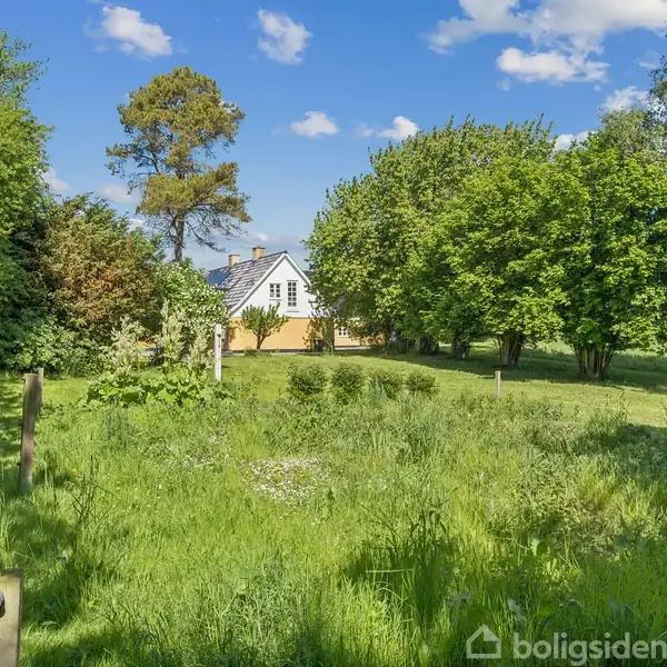 Et moderne køkken har hvide skabe og sort-hvidt ternet gulv. En stor ovn og emhætte er centralt placeret. Væggen er dækket med hvide fliser. Tavlen siger: "Velkommen Hjem Søde."