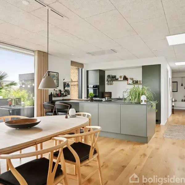 Et moderne køkken-alrum med træbord og stole, en køkkenø i midten og store vinduer, der viser en terrasse med planter. Lyse træ-gulve og hvidt loft danner rammen.