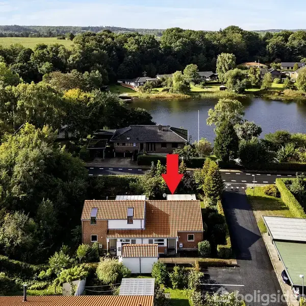 Et hus med rødt tag er i centrum markeret med en rød pil. Det ligger i et grønt område nær en sø og omgivet af andre huse og træer.