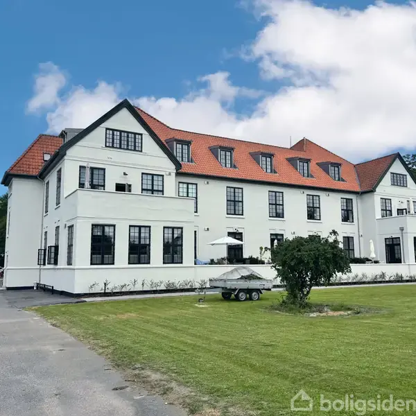 Stor hvid bygning med rødt tegltag står stille på en velplejet grøn græsplæne under en blå himmel med få skyer. Skilt med teksten "Bager" ses i baggrunden.