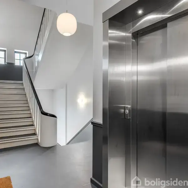 Elevator i et moderne opgangsrum, står stille ved siden af en trappe med sort rækværk. En rund loftslampe hænger ned fra loftet. Ingen tekst fundet.