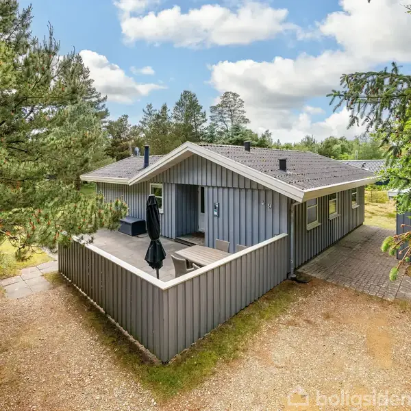 Et gråt træhus står stille omgivet af fyrretræer. Huset har en terrasse med en parasol. Himlen er let overskyet, hvilket skaber en fredelig atmosfære.
