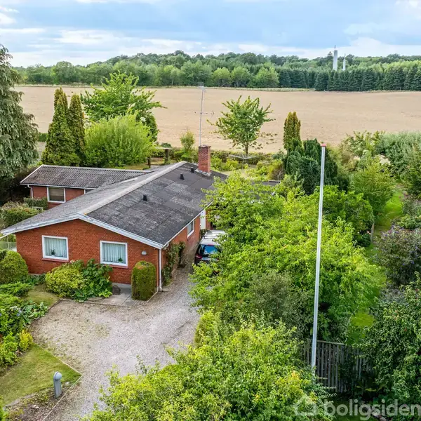 Et rødt murstenshus med sort tag ligger blandt frodig vegetation. En grusvej fører til huset, omgivet af træer og buske. I baggrunden ses en stor mark.