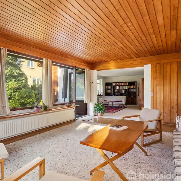 En stue med stribede sofaer og træstole står omkring et glasbord. Store vinduer med gardiner slipper lys ind, og en rund lampe hænger på væggen.