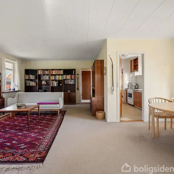 Stuen har et tæppe, sofa, sofabord, og reol fyldt med bøger. Et rundbord med stole står nær et vindue. Køkkenet er delvist synligt gennem en åbning.