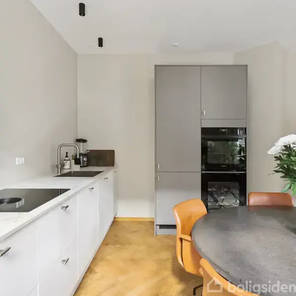 Et moderne køkken med hvide skabe og indbygget grydeholder ved siden af en vask. Et rundt spisebord med brune stole og en blomsterbuket skaber en hyggelig atmosfære.