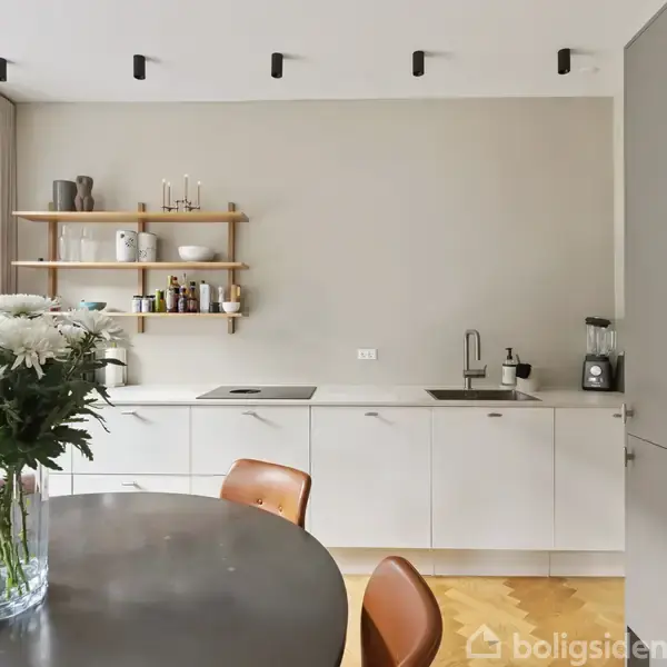 Et moderne køkken med hvide skabe; en glaskrukke med blomster på et rundt bord. Hylder med krydderier og kogebøger hænger over bordpladen. Rummet er lyst med trægulv.