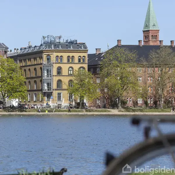 Cykel i forgrunden ved søbred; bagved ses historiske bygninger med en kirke og grønne træer, der skaber en rolig bylandskab.