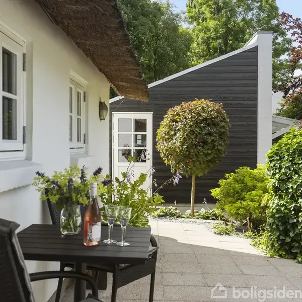 En terrasse med et lille bord og to stole, hvor en vinflaske og glas står, omgivet af en hvid murstensbygning og grønne buske i en have.