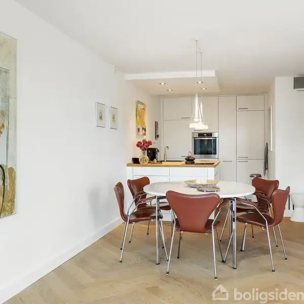 Et rundt spisebord er omgivet af seks brune stole i en lys, moderne spisestue. Et stort maleri hænger på væggen, og et åbent køkken ses i baggrunden.