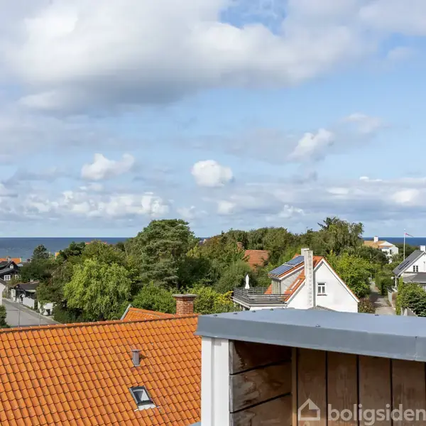 Røde tagtage strækker sig mod horisonten, mens grønne træer omgiver huse under en blå himmel med skyer. I det fjerne ses havet.