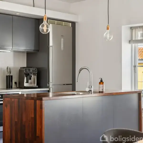 Køkken med mørkt træbord er udstyret med en kaffemaskine og moderne apparater. En stor vindue lader lys strømme ind. Et simpelt indrettet rum med hængende lamper.