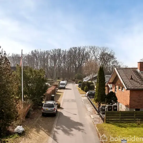 En stille villavej med huse på begge sider. Flere biler er parkeret langs vejen. Baggrunden domineres af en skov med træer uden blade under en blå, overskyet himmel.