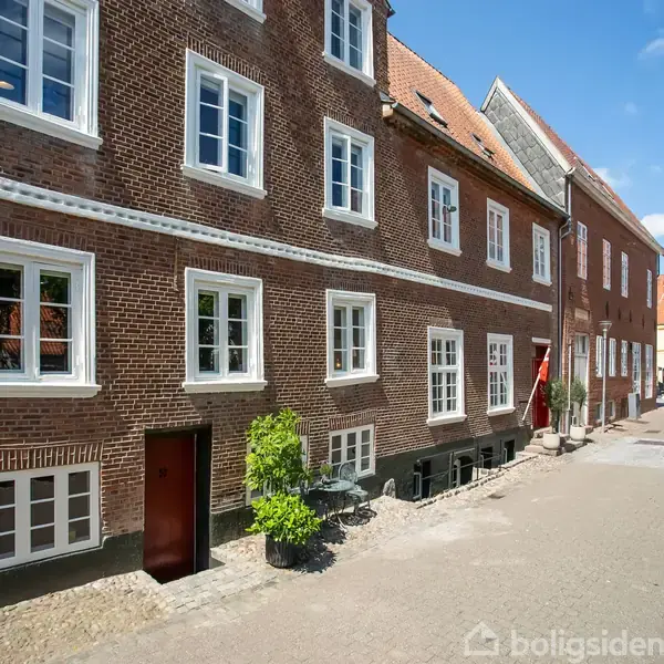Brun murstensbygning med hvide vinduer flankerer en brostensbelagt gade. Folk går i baggrunden under en blå himmel med få skyer. Hoveddøren er placeret nær bunden af bygningen.