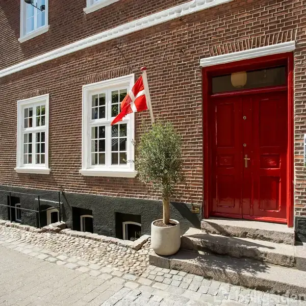 Rød dør prydet med en dansk flag i en murstensbygning. To træer i krukker flankerer indgangen. Omgivet af brostensbelagt gade og flere vinduer. Adresseplade viser "1".
