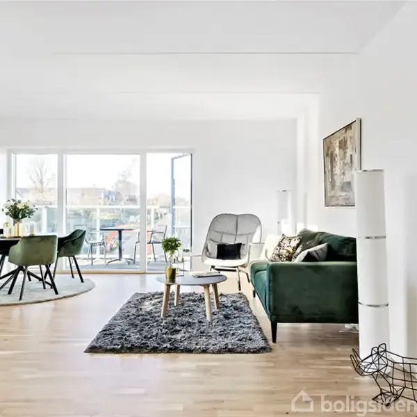 Stue med grøn sofa og sort sofabord på gråt tæppe, ved siden af stor gulvlampe. Spisebord med stole ved vinduesparti. Væggene er hvide, gulvet er i lyst træ.