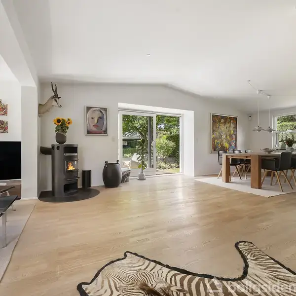 Stue med træbrændeovn omgivet af moderne møbler; zebra-tæppe spredt på trægulv. Store vinduer slipper naturligt lys ind og viser have udenfor. Dekorative vægmalerier og en vase med blomster.