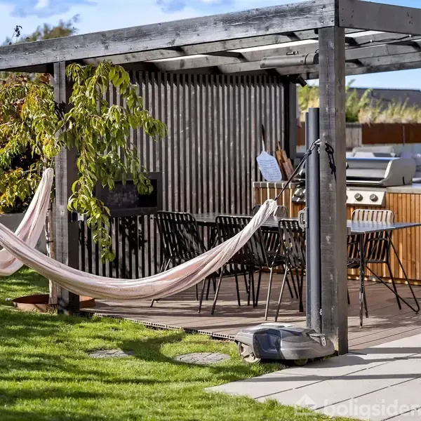 Hængekøje hænger under en pergola ved et udendørs spiseområde med bord og stole. Omgivelserne inkluderer en grøn have, grill og udendørs køleskab.