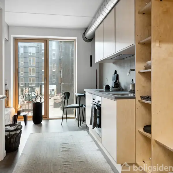 Et smalt køkken med en ovn og vask på siden, oplyst af dagslys fra glasdøre til en altan. Ved siden af dørene er der placeret en stol.