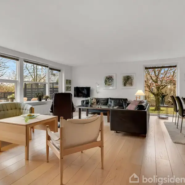 Stue med sofa og stole (objekt) arrangeret omkring et sofabord (handling). Store vinduer og døre slipper lys ind (kontekst). Spiseområde med bord og stole i baggrunden.