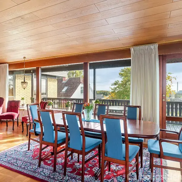 Spisebord med otte blåstole står på et rødt tæppe i en stue med trægulv og stort vinduesparti. Røde lænestole er arrangeret i hjørnet ved siden af et lille bord.