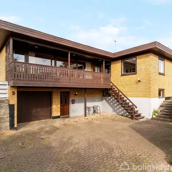 Et étplanshus i gul mursten med en bred balkon og trætrappe. Det har en garage og en delvist brolagt indkørsel omgivet af træer og buske. Husnummeret er 9.