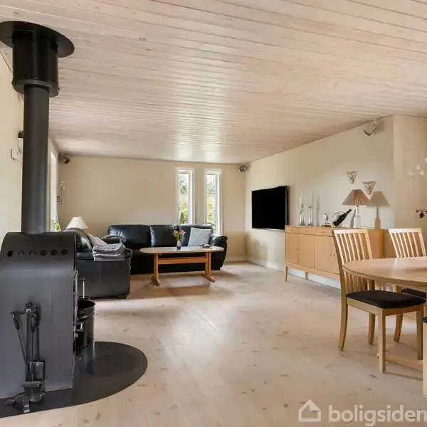 Brændeovn står til venstre, tilfører varme i stuen med lyst trægulv, sofa og sofabord til højre, spisebord med blomster til højre, fladskærms-tv på væggen.