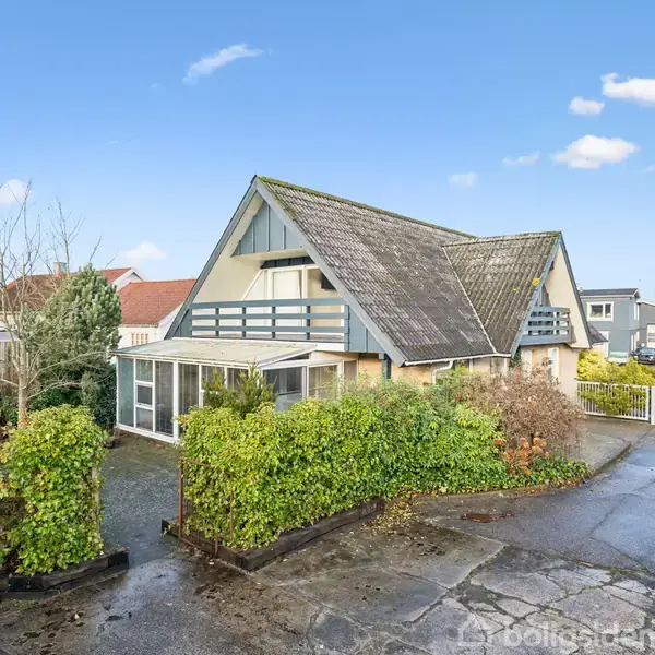 Et hus med skråt tag står blandt buske og træer på en villavej. I forgrunden ses en flisebelagt indkørsel og omgivende grøn beplantning under en blå himmel.