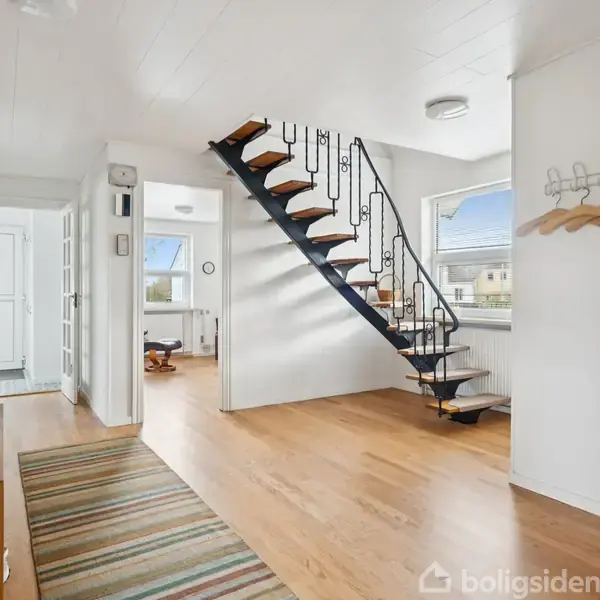 Trappe med trætrin og sort gelænder fører opad i en lys entre. Væggene er hvide med et stribet tæppe på gulvet. Vindue til venstre for trappen.