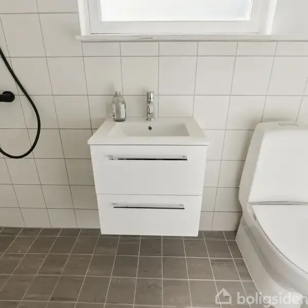 En hvid håndvask med to skuffer står fast på en flisevæg ved siden af et toilet. Et brusehoved hænger til venstre i det flisebelagte badeværelse.