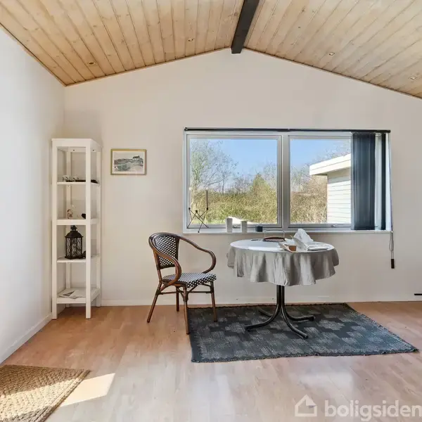 Et rundt bord med dug og kaffekopper står ved et vindue. En flettet stol er ved bordet. Værelset har lyst træloft og trægulv med et tæppe.