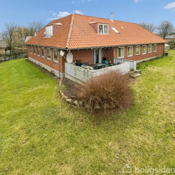 En stor rød murstensbygning med skråt tag står på en grøn plæne. Bygningen har en terrasse med havemøbler, omgivet af åben have og træer under en klar blå himmel.