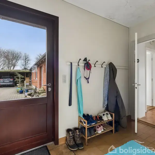 Dør åbner ud mod indkørsel med træer. Indendørs ses en gang med skohylde og tøjknager til jakker og tørklæder. En åben dør fører videre til andre rum.