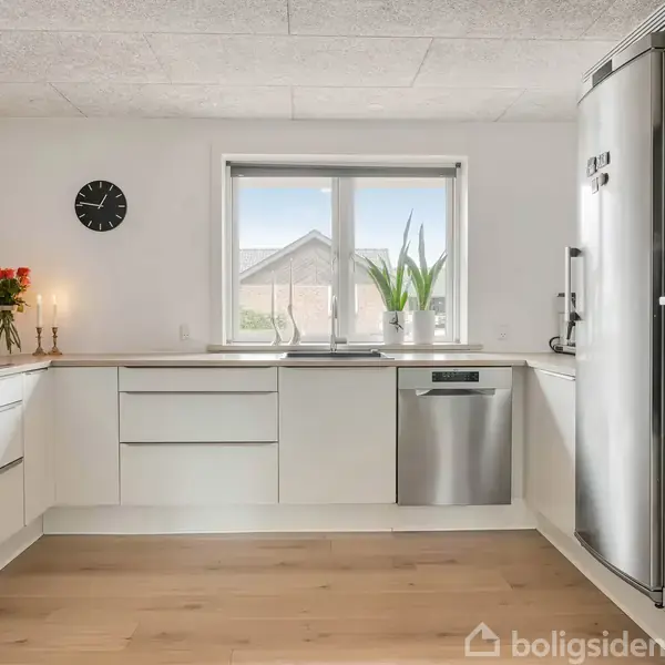 Et moderne køkken med hvide skabe og stålapparater som køleskab og ovn. På bordpladen står stearinlys og en vase med blomster. Et vindue viser udsigt til et hus. Der er et ur på væggen.