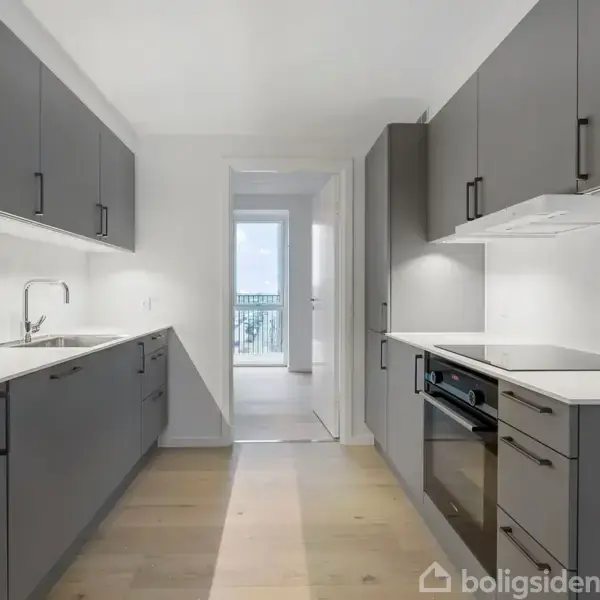Moderne køkken i mørkegrå farver, har indbyggede skabe og apparater og en lys trægulv. En vask og kogeplade er synlige. En lysåben dør fører til en balkon.