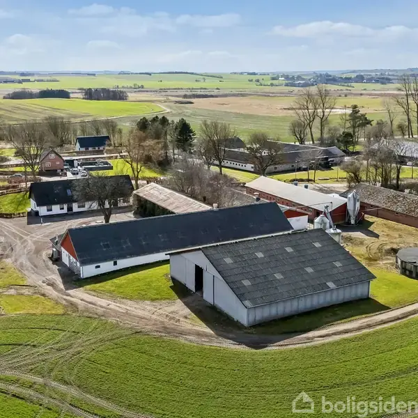 Landbrugsbygning med sort tag ligger i et åbent landbrugsområde omgivet af marker og flere mindre bygninger. Landskabet strækker sig under en blå himmel med spredte skyer.
