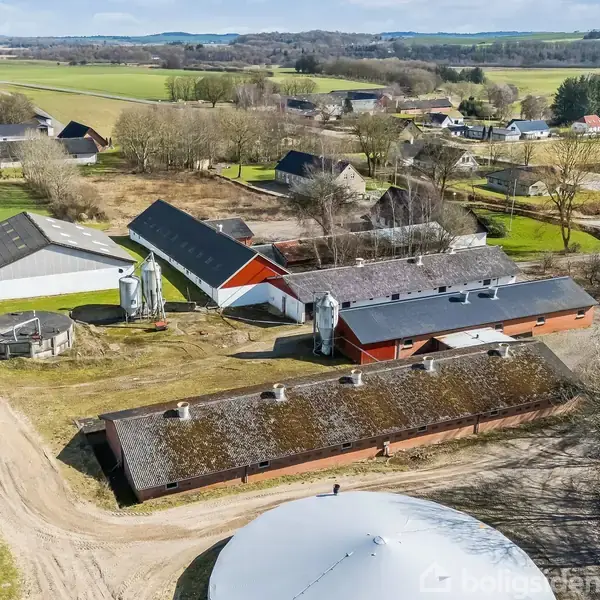 Landbrugsbygninger ligger spredt i et grønt landskab med marker og træer. Et beboelsesområde med huse ses i baggrunden. Det er en lys dag med klar himmel.