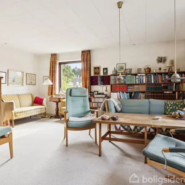 Stue med sofagruppe bestående af lyseblå møbler omkring et træbord. Bøger fylder reoler ved væggen. Store vinduer med gardiner lader lys ind, skaber en lys og indbydende atmosfære.