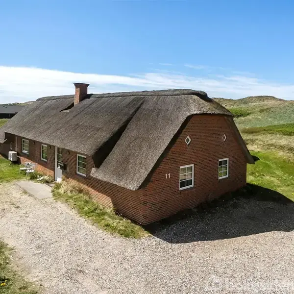 Et rødt murstenshus med stråtag står stille i et åbent, grønt landskab med klare himmel. Nummer "11" ses på gavlen. Grusvej og sandklitter omgiver bygningen.