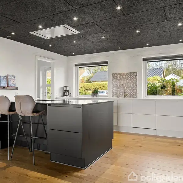 Et moderne køkken med en sort køkkenø og to barstole. Køkkenet er lyst med store vinduer, trægulv og hvidmalede vægge. En kaffemaskine og planter står i vinduet.