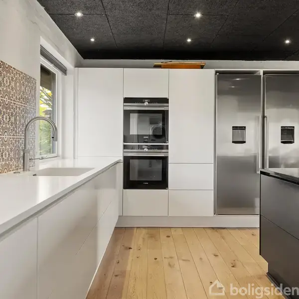 Et moderne køkken med hvide skabe og indbyggede ovne til venstre. Ved siden af er der et stort, rustfrit stålkøleskab. Trægulv og sort loft med indbyggede spots.