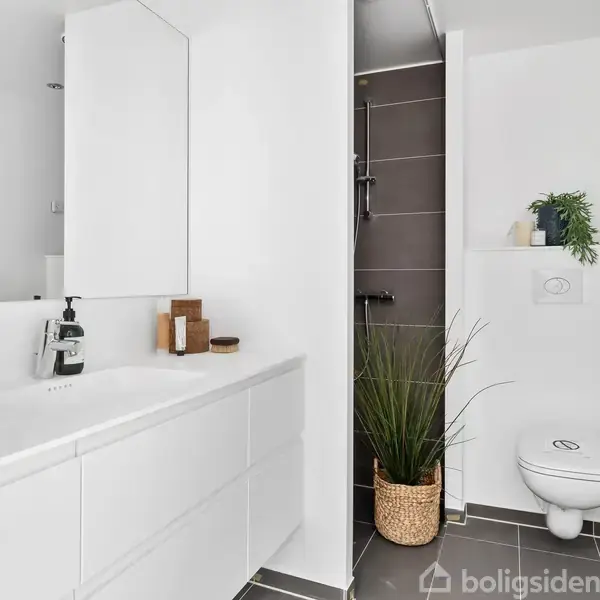 Hvidt badeværelse med væghængt toilet og møbler, bruseområde med mørke fliser, plante i kurv ved siden af toilet, og håndvask med sæbe og dekorationer.